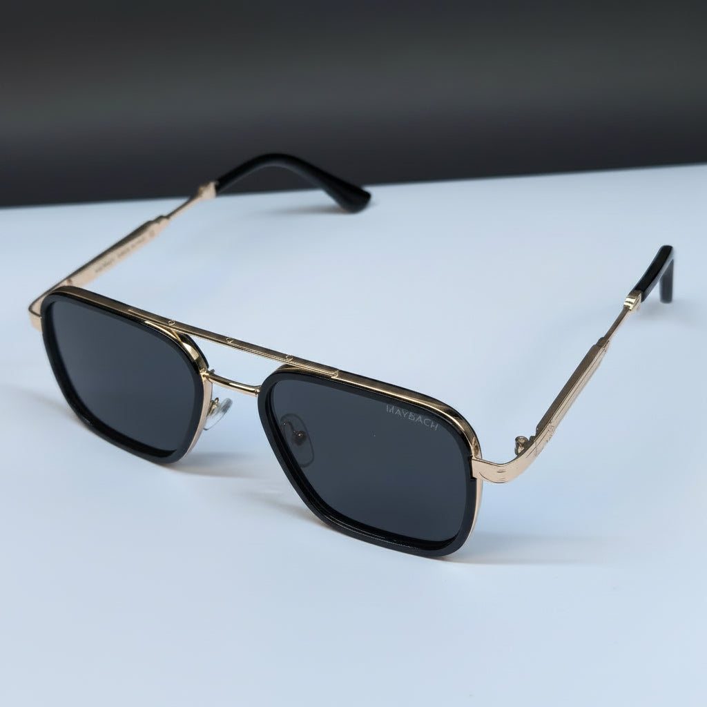 A pair of square aviator-style sunglasses with gold-tone metal frames, dark lenses, and black temple tips, resting on a white surface with a dark gray background.